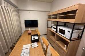 A room with a long wooden table, chairs, a flat-screen TV, and built-in shelves with appliances. - Core Royal-Vacation Rental (Kyoto)