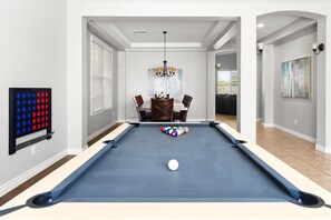 A pool table in a room with a dining area, a chandelier, and a painting on the wall.