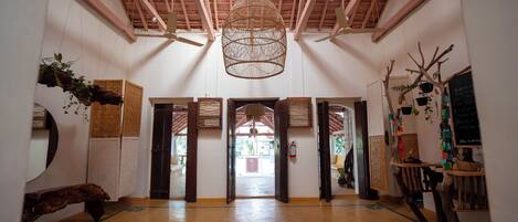 Un hall d’entrée spacieux avec un plafond haut, des poutres en bois et un lustre suspendu en osier. Plusieurs portes donnent accès à d’autres espaces, et le sol présente un motif géométrique.