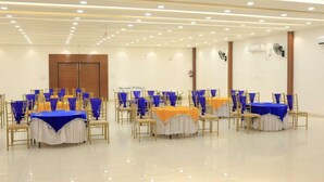 A spacious hall with multiple round tables, each covered with blue and orange tablecloths, and gold chairs with blue seat covers. - Evana Rajaji  (Ganga Bhogpur Malla)
