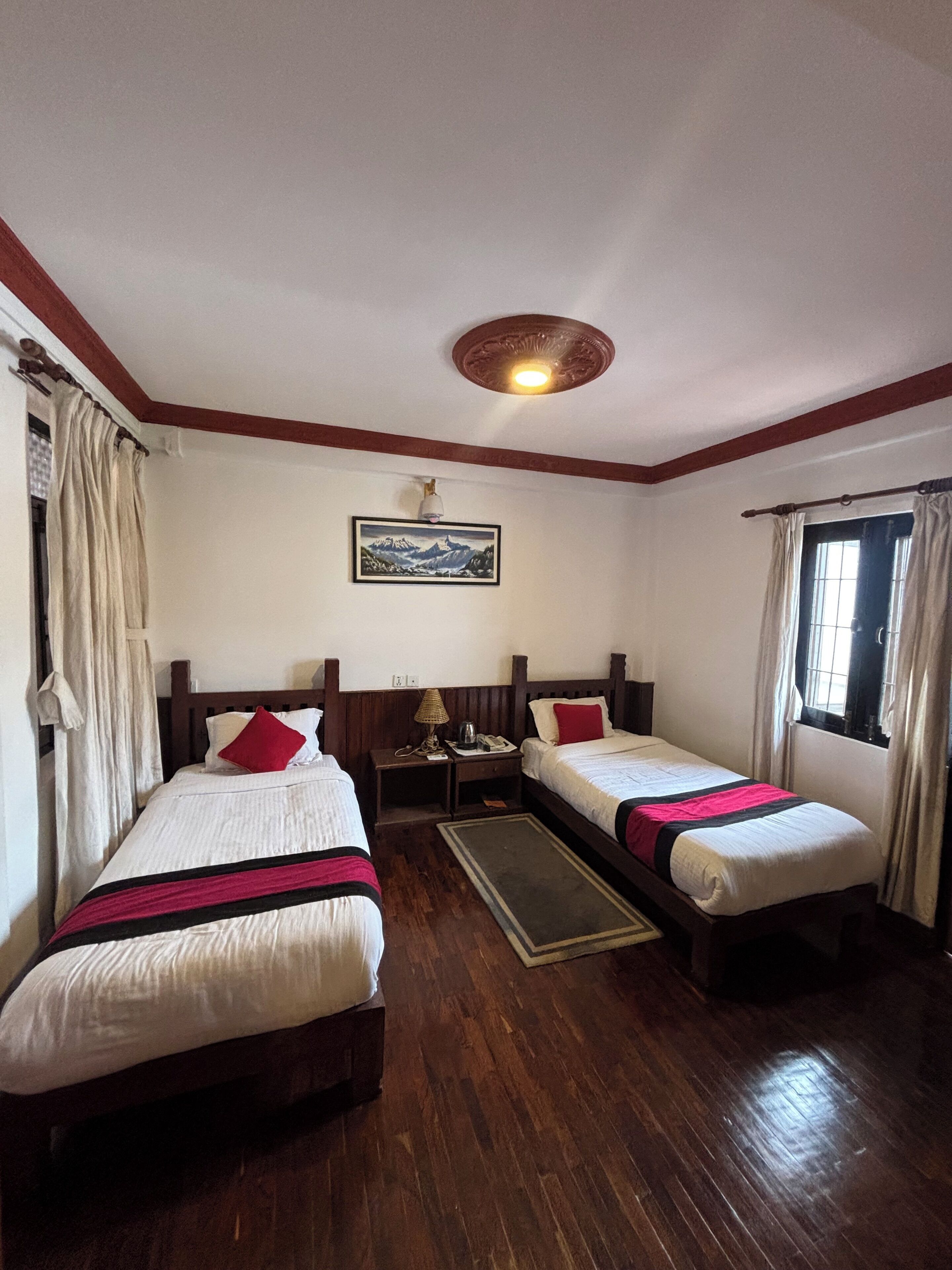A hotel room with two single beds, wooden flooring, a ceiling light fixture, a painting on the wall, and a window with curtains.