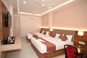 A hotel room with a wooden desk, two beds, a ceiling fan, and a wall-mounted TV. - Hotel Nirvvana Residency (Varanasi)