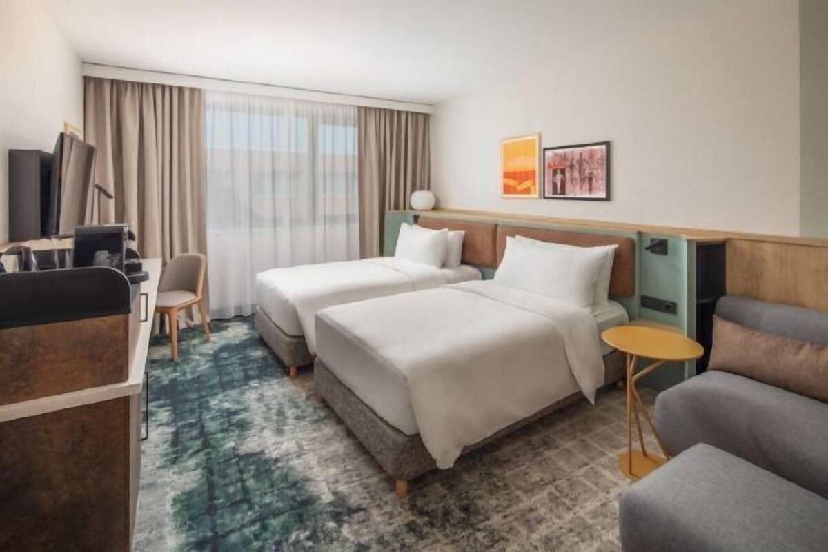 A hotel room with two beds, a desk, a chair, a small table, and a large window with curtains.