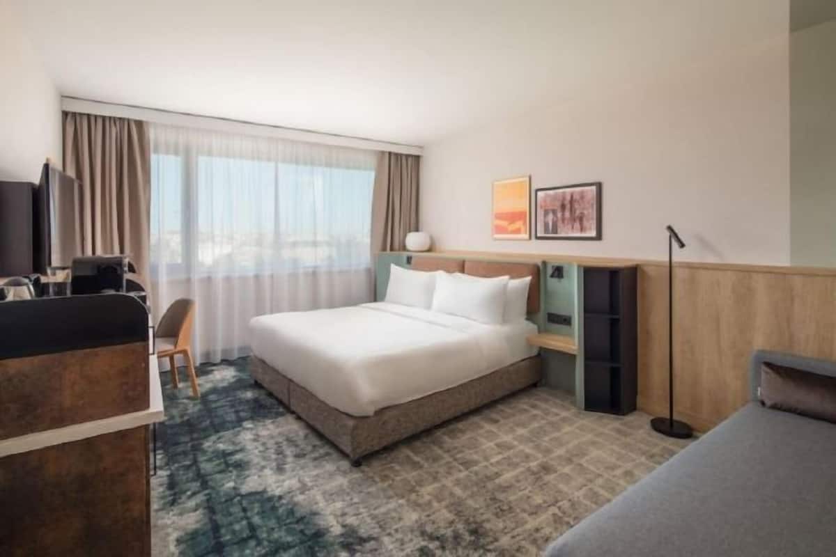 A hotel room with a bed, a desk, a chair, a TV, and a large window with curtains.