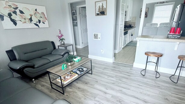 Un salon avec un canapé gris, une table basse avec des magazines, un ventilateur de plafond et un coin cuisine doté d’un comptoir et de tabourets de bar.