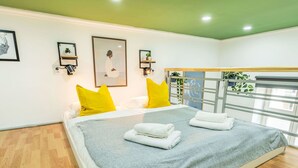 Una habitación de hotel moderna con una cama grande, almohadas amarillo brillante, una manta gris y toallas cuidadosamente dobladas. Hay cuadros en la pared y un balcón con barandilla y plantas.