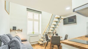 Un espacio habitable moderno con un sofá gris, piso de madera, un área de comedor con sillas, una escalera y un televisor montado en la pared.