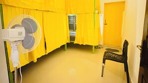 Een kamer met gele gordijnen, een staande ventilator, een stoel en een deur.