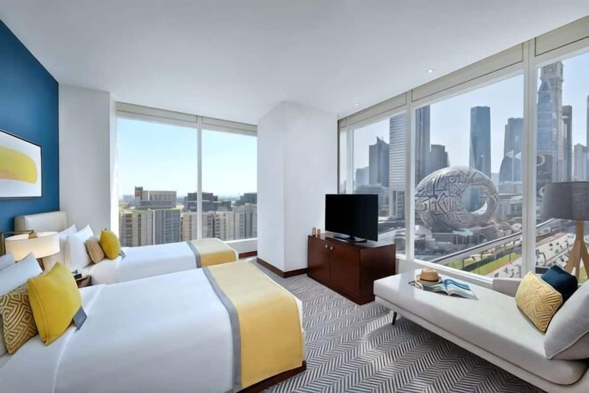 A hotel room with two beds, a television, a sofa, and a large window with a city view.