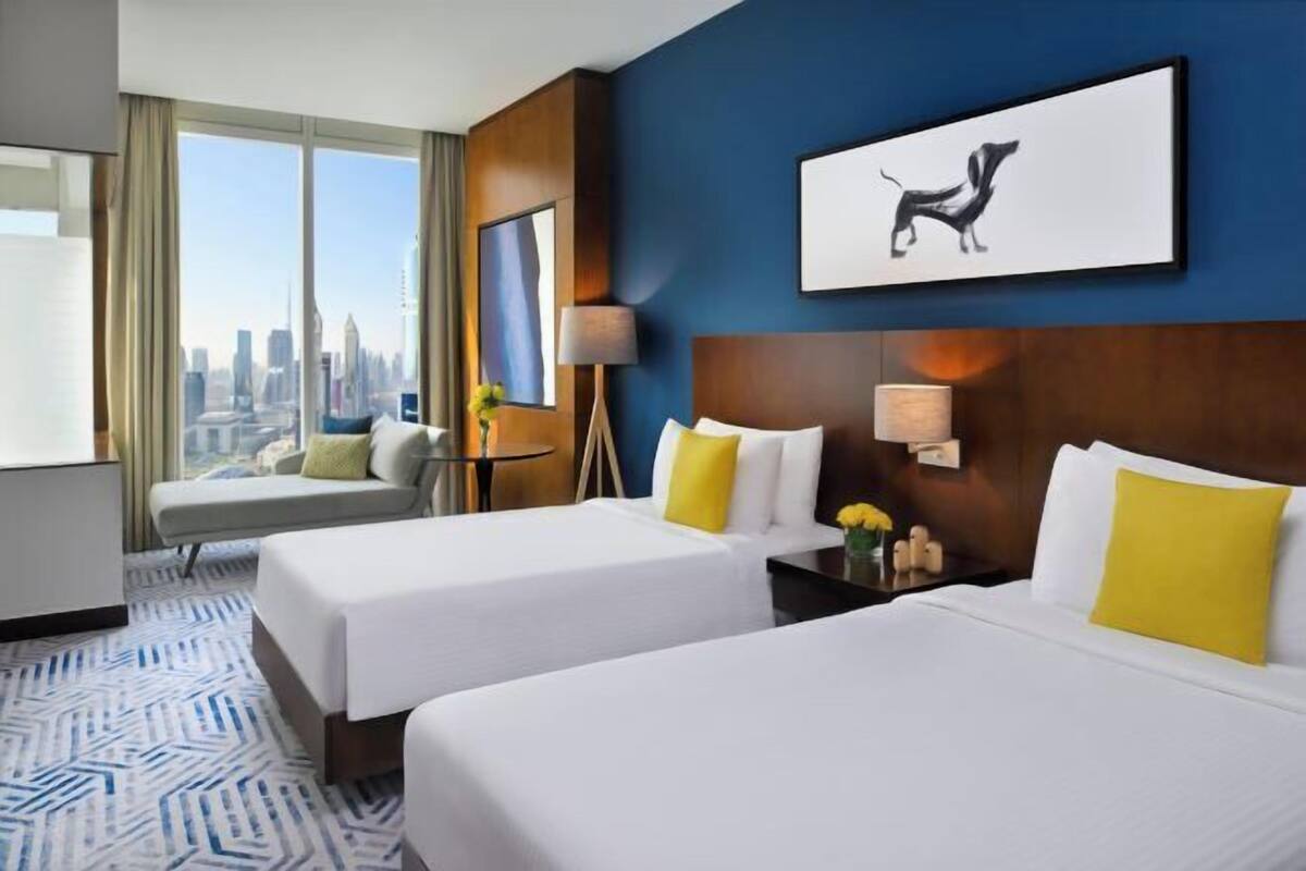 A hotel room with two beds, a city view, a framed artwork, and a desk with a lamp.