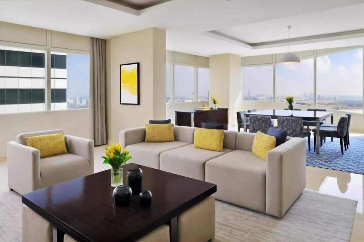 A modern hotel room with a sofa set, a dining table, and a view of the cityscape.