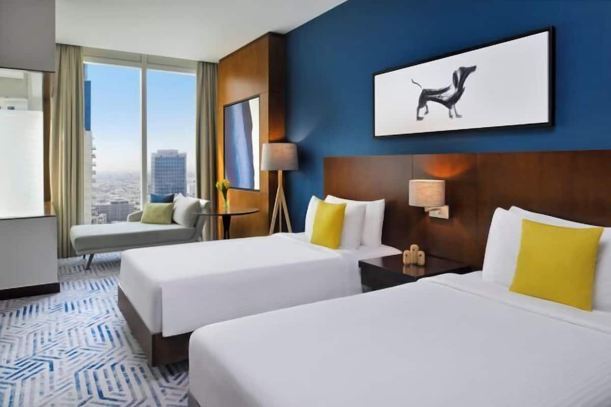 A hotel room with two beds, a city view, a framed artwork, and a desk with a lamp.
