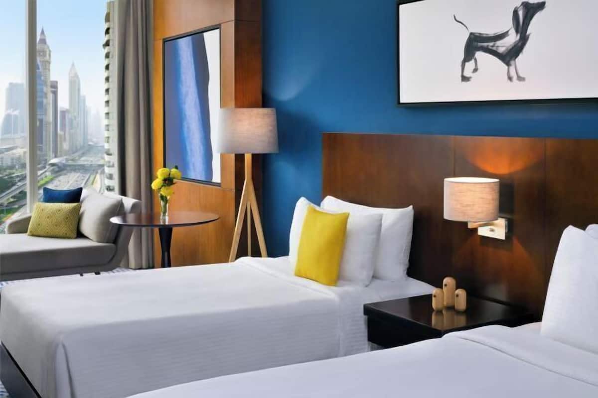 A hotel room with two beds, a view of a cityscape, a painting of a dog, and a lamp on the nightstand.