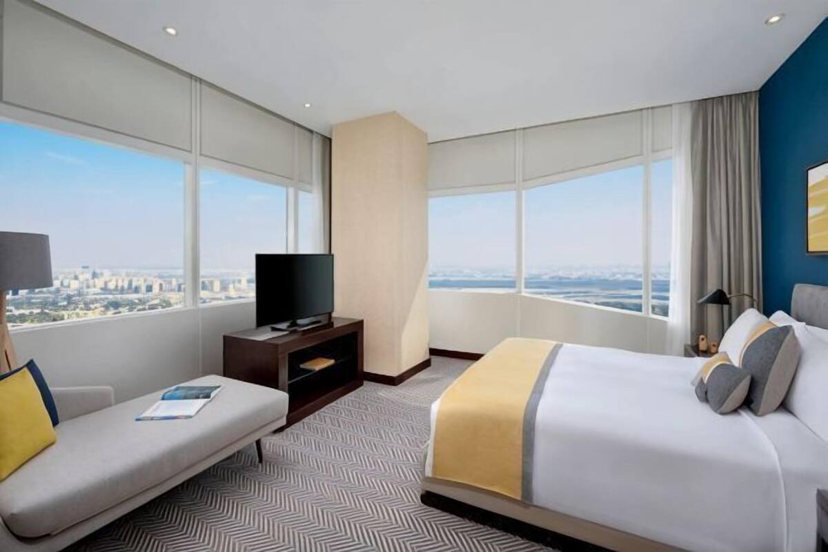 A hotel room with a bed, a sofa, a television, and a large window offering a city view.