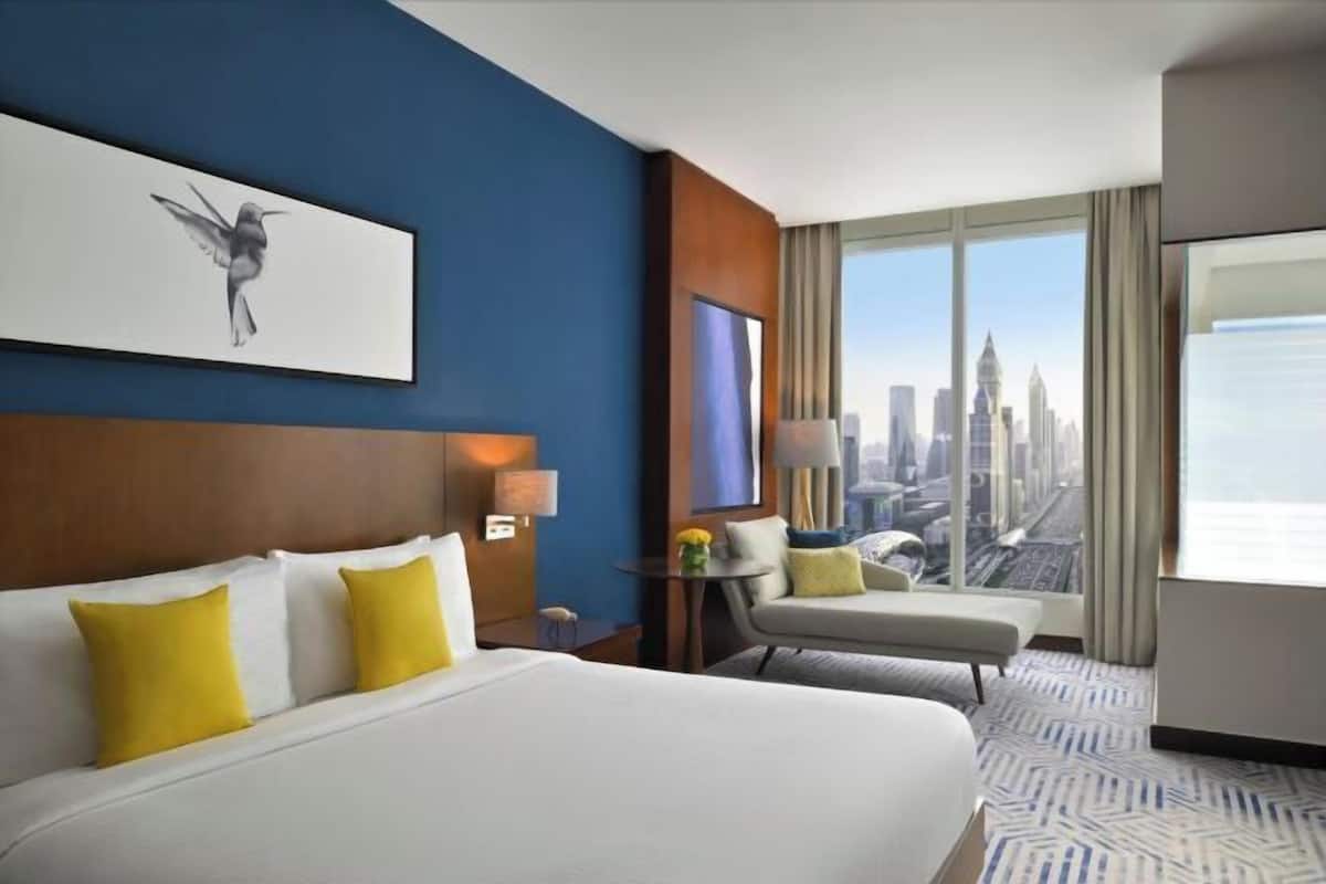 A hotel room with a bed, a chair, a small table, a lamp, and a city view.