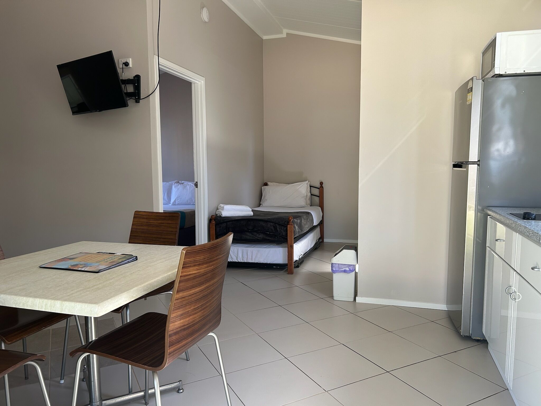 A compact hotel room with a bed, a small kitchenette, a dining area with a table and chairs, and a TV mounted on the wall.