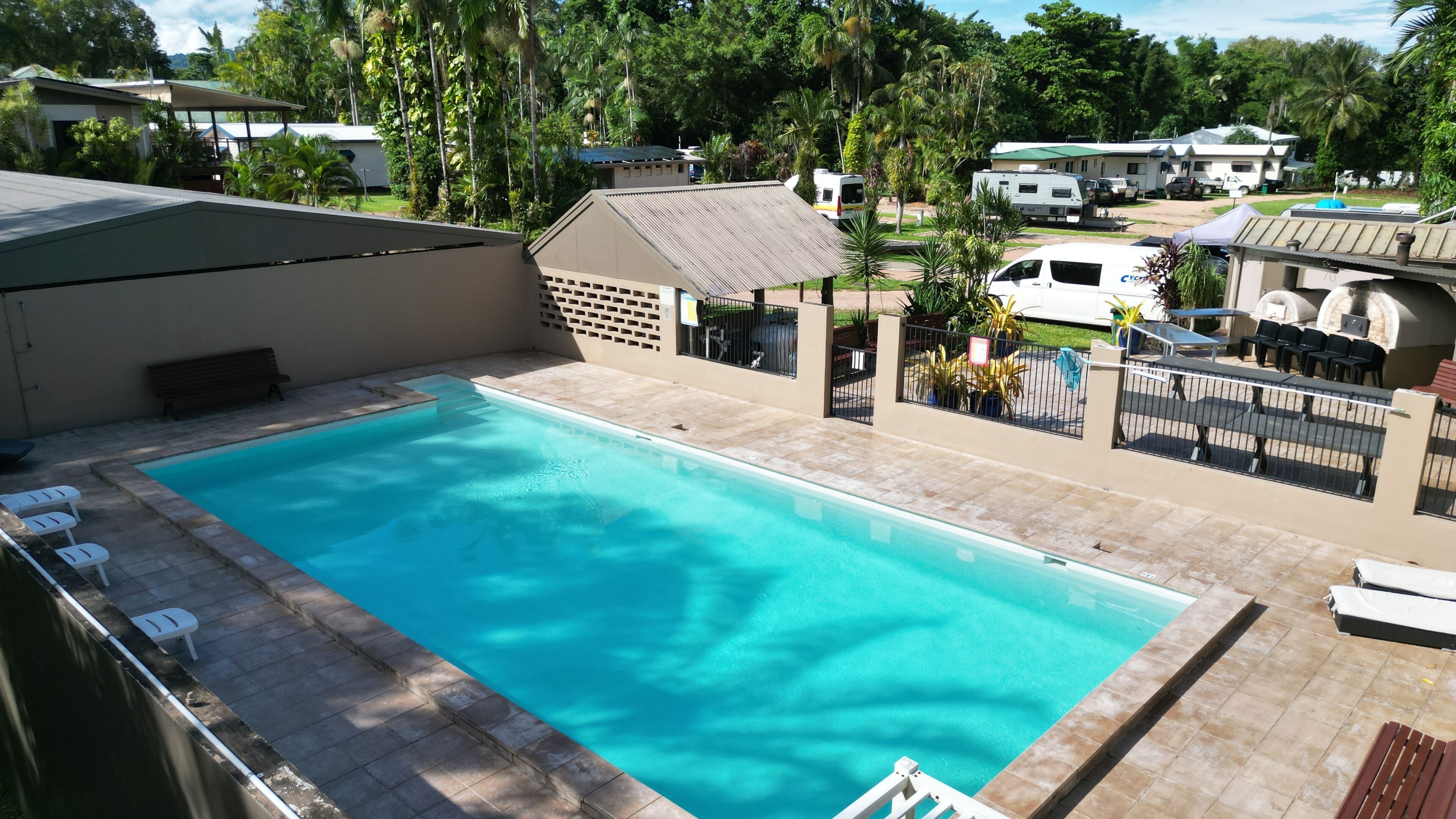 A swimming pool with a tiled deck, a fenced area with outdoor seating, and a view of a residential area with houses and RVs in the background.