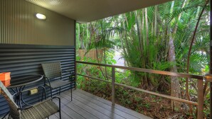 Un balcón con sillas de mimbre y una mesa, rodeado por una barandilla de metal, con vista a un exuberante jardín tropical.