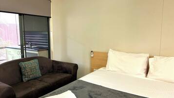 Quarto de hotel com cama, sofá, janela com cortinas e uma pequena cozinha.