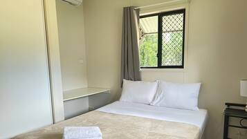 Yhden hengen sänky, jossa on beigenvärinen peitto, valkoinen tyyny ja taitettu valkoinen pyyhe. Ikkunassa on metallinen ritilä, ja seinällä on pieni pöytä, jossa on lamppu, sekä pistorasia.
