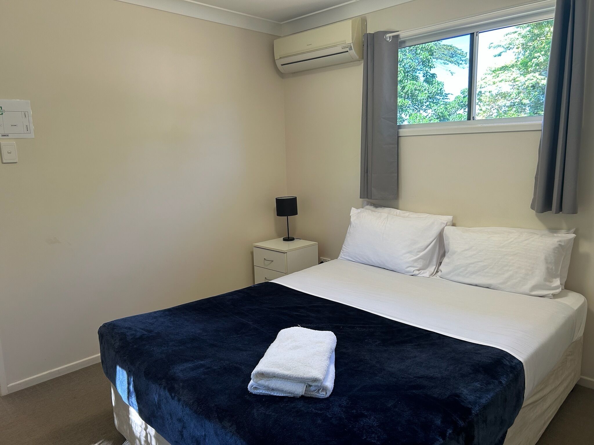 A bedroom with a bed, a nightstand, an air conditioning unit, and a window with curtains.