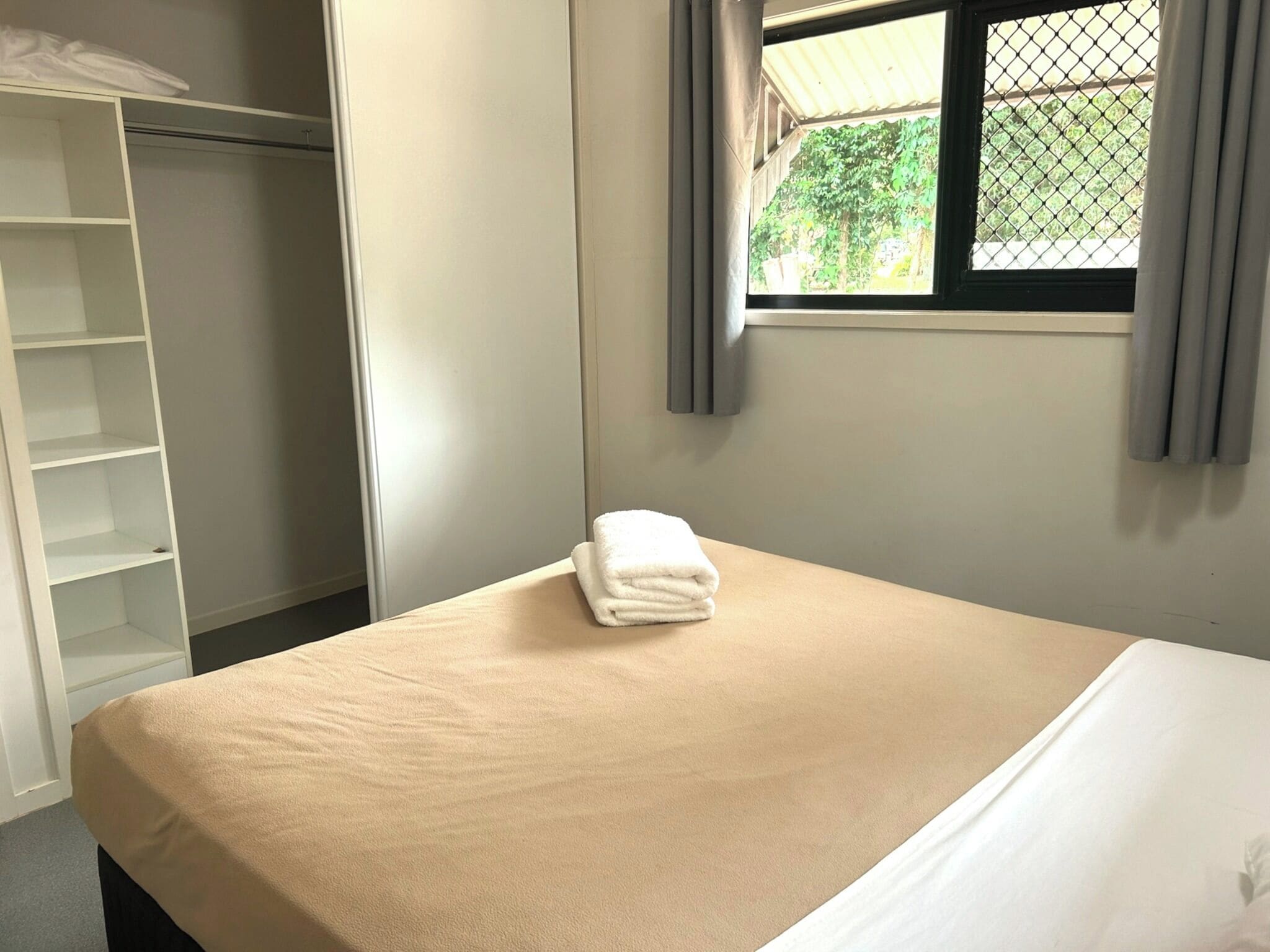 Un lit simple avec une couvre-lit beige et une serviette blanche pliée, une armoire blanche et une fenêtre donnant sur un paysage verdoyant.