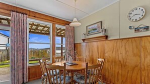 A dining area with a wooden table and chairs, a clock on the wall, and a view of the outdoors through large windows. - Sunny Hilltop Red Bach w Lake and Mountain Views (Taupō)