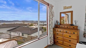 A bedroom with a large window, a wooden dresser, a mirror, and a 'Home' sign on the wall. - Sunny Hilltop Red Bach w Lake and Mountain Views (Taupō)