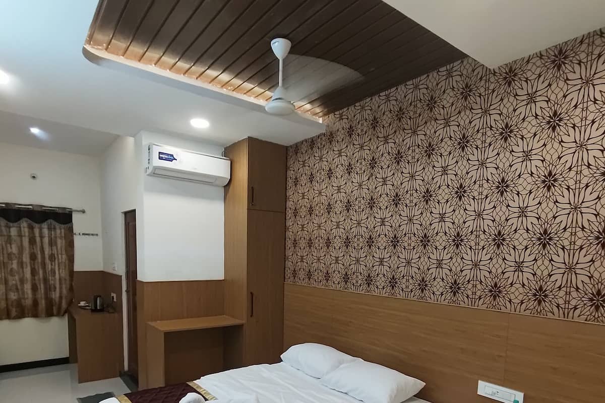 A modern hotel room with a large bed, a ceiling fan, an air conditioning unit, a patterned wall, and a small bedside table with a bottle of water.