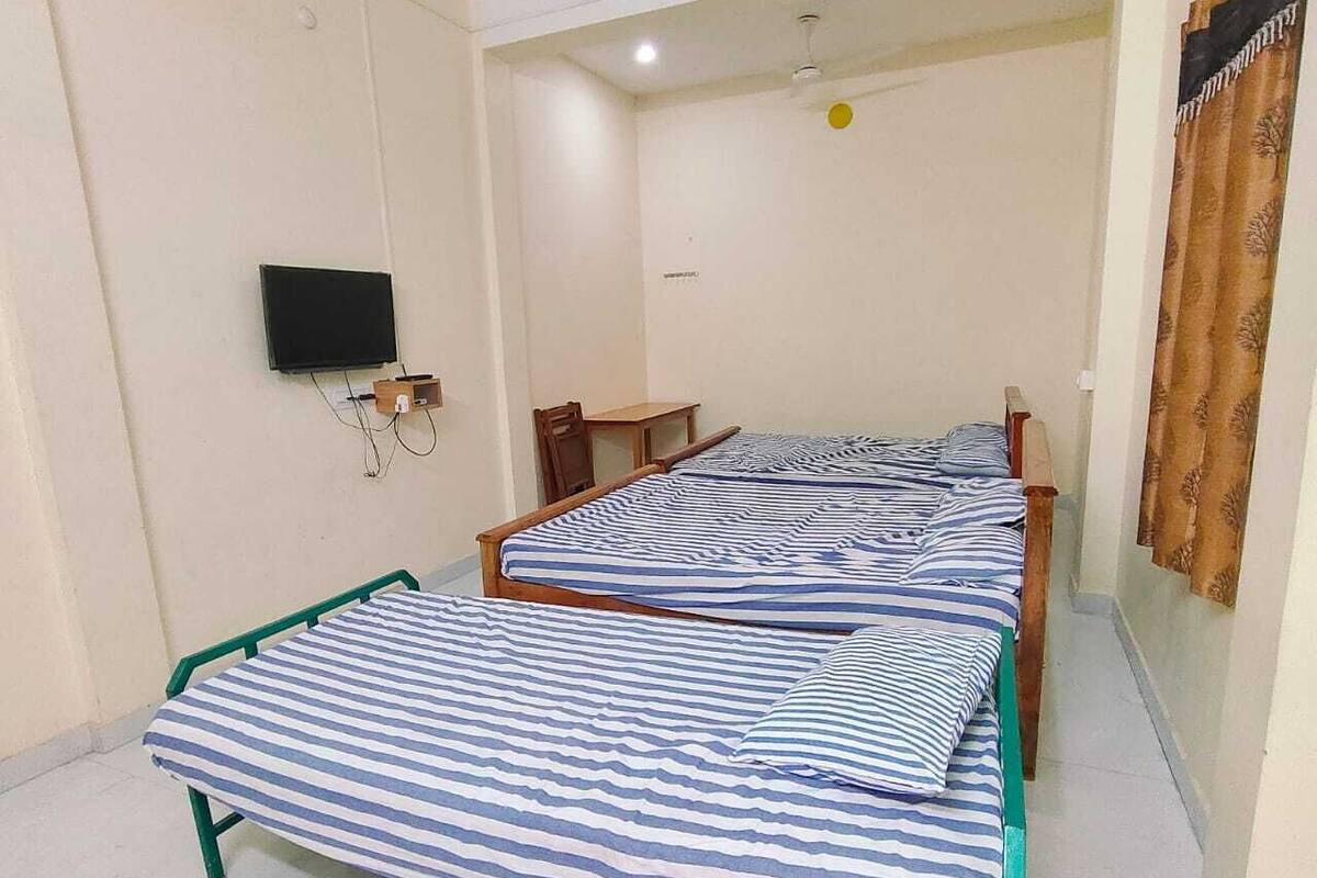 A room with two single beds, a small table, a chair, a television mounted on the wall, and a ceiling fan.