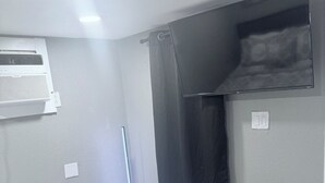 A bedroom with a bed, an air conditioning unit, a TV mounted on the wall, and a window with a curtain.