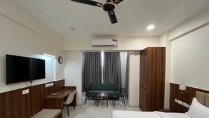 A hotel room with a bed, a desk, a chair, a sofa, a TV, an air conditioning unit, a ceiling fan, and curtains. - KRISHNA ANANDAM PALACE (Mathura)