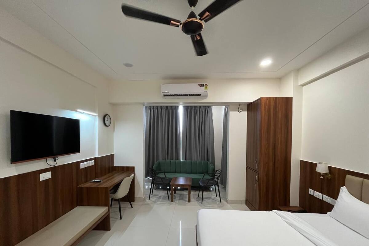 A hotel room with a bed, a desk, a chair, a sofa, a TV, an air conditioning unit, a ceiling fan, and curtains.