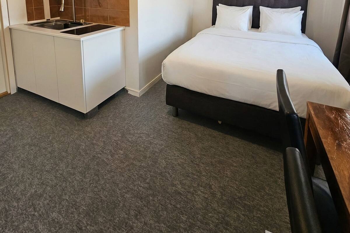 Une chambre d’hôtel compacte, dotée d’une kitchenette, d’un lit avec une tête de lit sombre et d’une seule chaise.