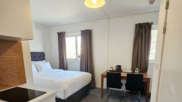 A hotel room with a bed, a desk, a chair, a small kitchenette, and a window with curtains. - Nox Hotel (Huddinge)