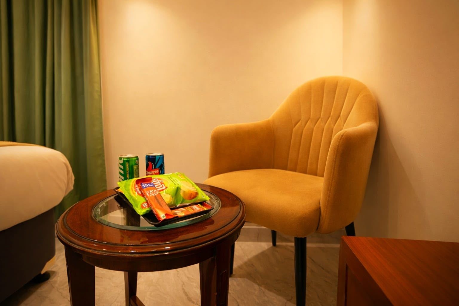 Habitación de hotel con un sillón amarillo, una mesita de vidrio con aperitivos y una cama.