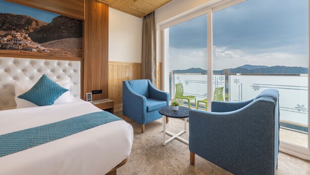 A hotel room with a large bed, a blue armchair, a small table, and a view of the mountains through a large window. - The Grand Welcome Hotel  (Shimla)