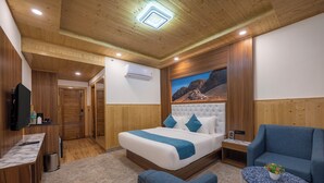 A hotel room with a wooden ceiling, a large bed with blue bedding, a blue armchair, a small table with a plant, and a TV on a wooden cabinet. - The Grand Welcome Hotel  (Shimla)