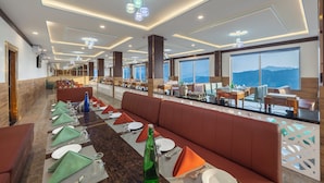 A modern hotel lobby with a long dining table, comfortable seating, and large windows offering a view of the mountains. - The Grand Welcome Hotel  (Shimla)
