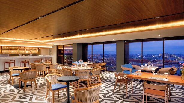 A modern dining area with tables and chairs, large windows with a city view, and a wooden ceiling. - Oakwood Yogyakarta (Yogyakarta)