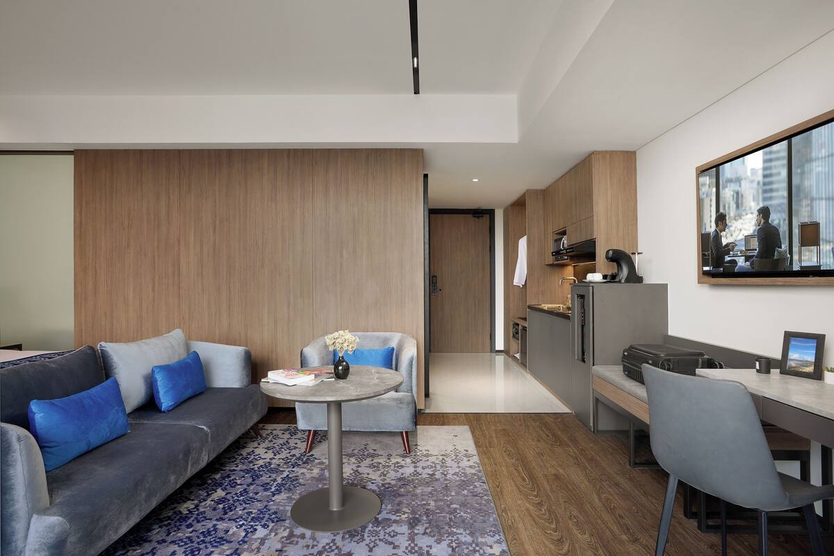 A modern living room with a grey sofa, a small round table, and a blue armchair. There is a kitchen area with a refrigerator, a sink, and a window overlooking the city.