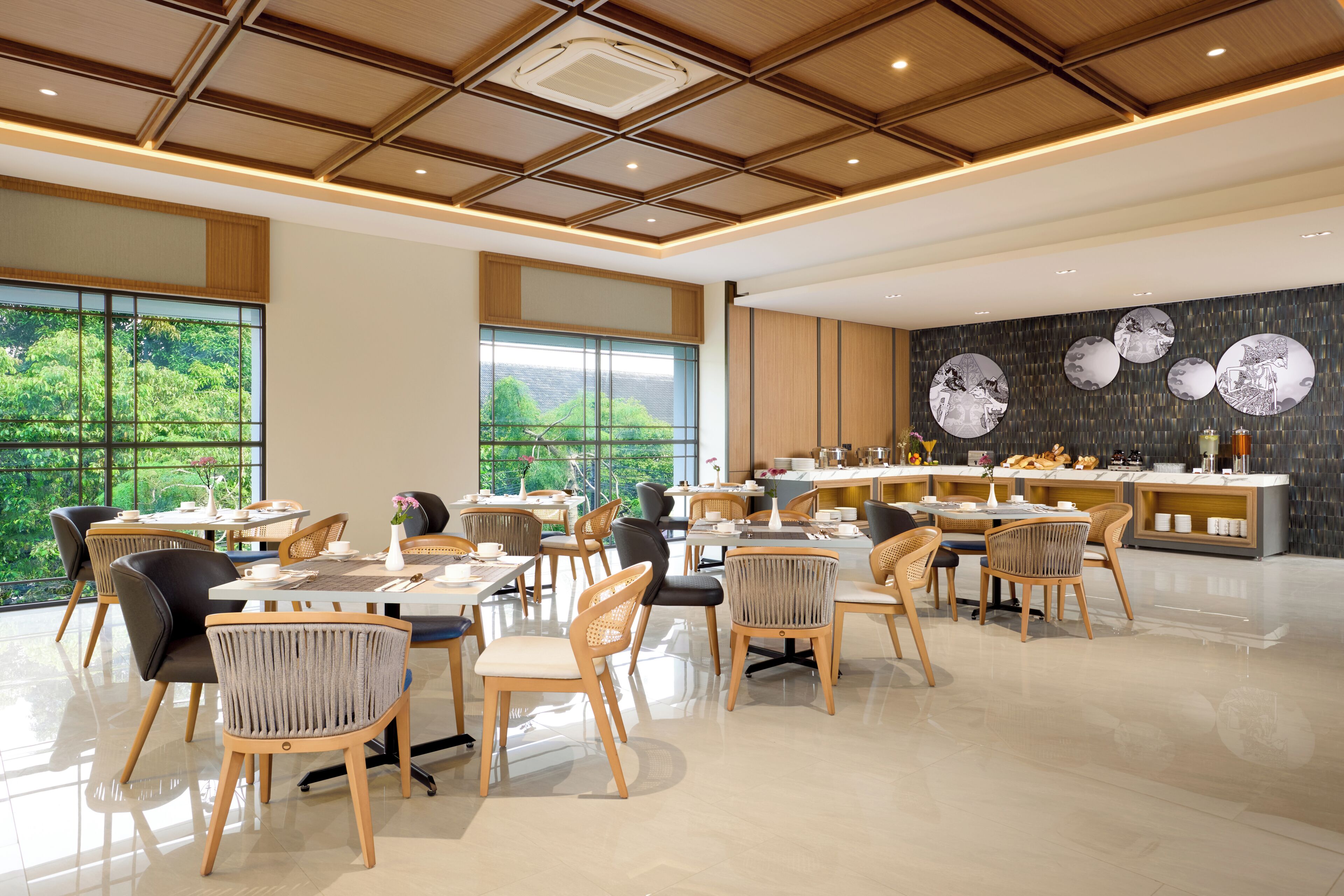 A modern dining area with multiple tables and chairs, large windows offering a view of greenery, and a kitchen area with a counter and decorative wall art.