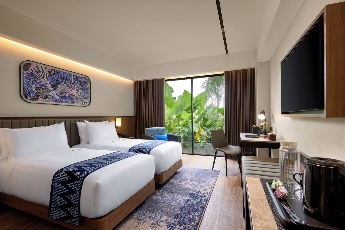A hotel room with a large bed, a desk, a chair, a TV, and a view of greenery outside.