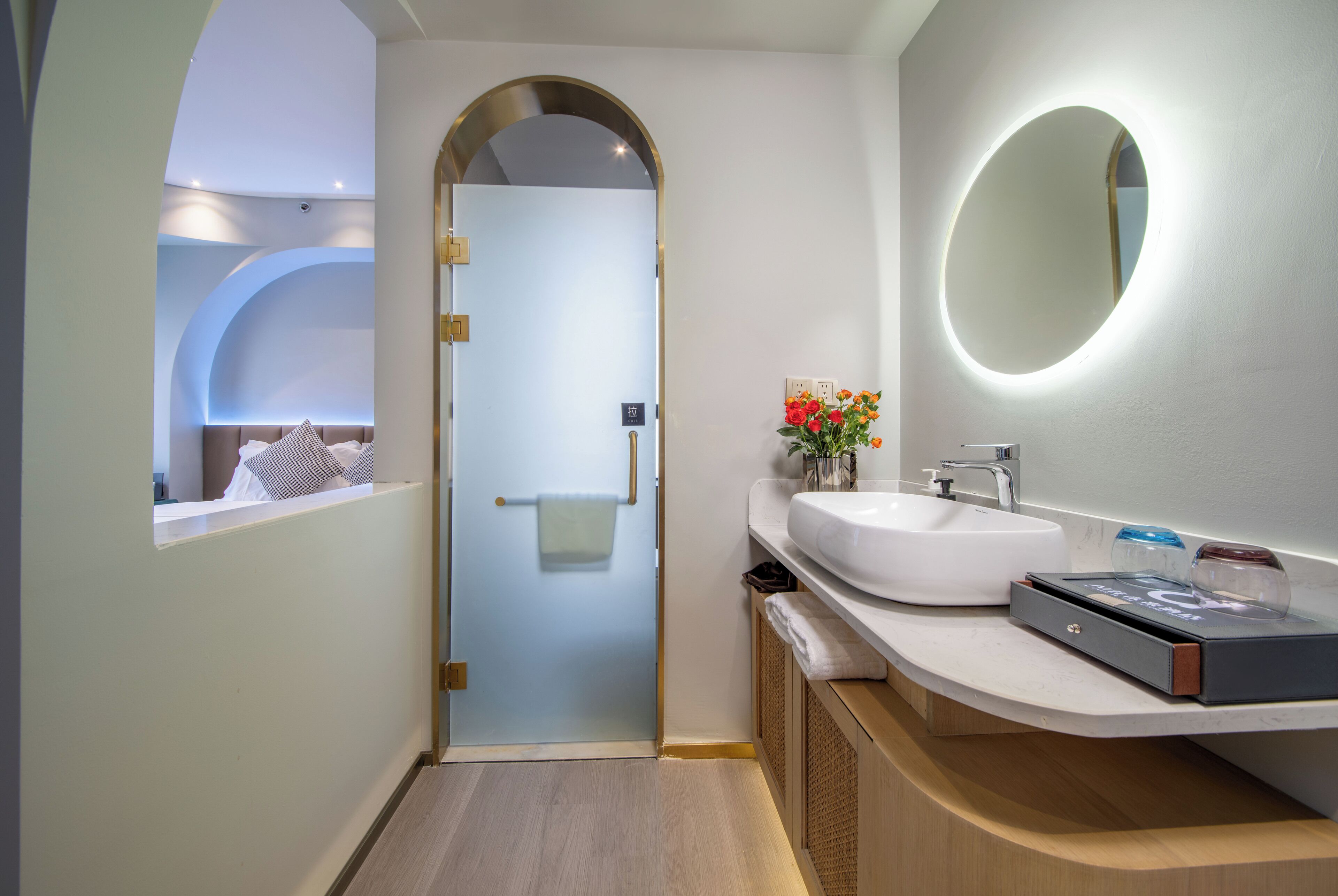 A modern bathroom with a glass door, a round mirror, a white sink, and a small vase with flowers.