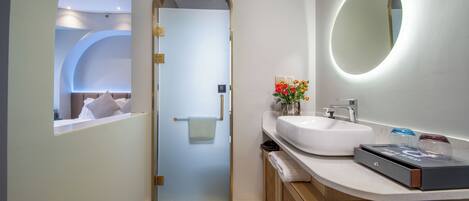 A modern bathroom with a glass door, a round mirror, a white sink, and a small vase with flowers.