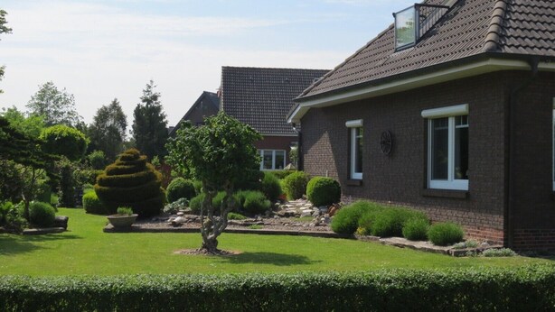 Een woonhuis met een schuin dak, een zichtbare schoorsteen en een verzorgde tuin met diverse struiken en gazon.