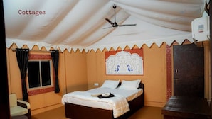 Habitación individual con cama, ventilador de techo, ventana con cortinas, cabecera decorativa y una mesita pequeña.