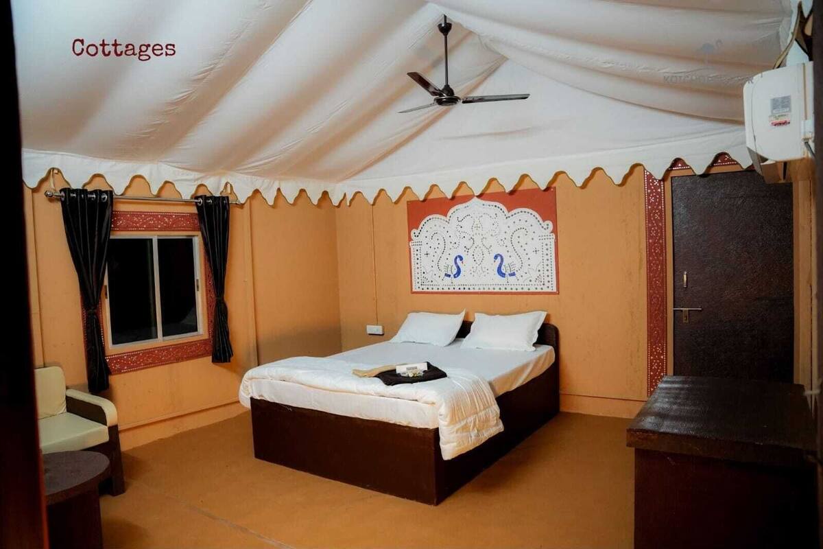 A single-bed room with a ceiling fan, a window with curtains, a decorative headboard, and a small table.