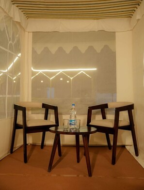 A Rajwadi Tent sit-out area with two wooden chairs and a small round table, a bottle of water, and a glass. - The Desert Resort (Bhuj)
