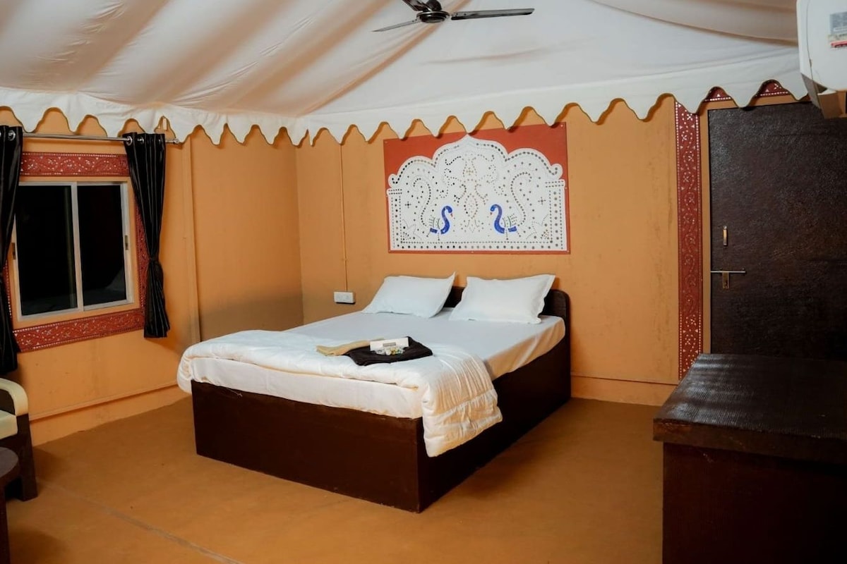 A single-bed room with a ceiling fan, a window with curtains, a decorative headboard, and a small table.